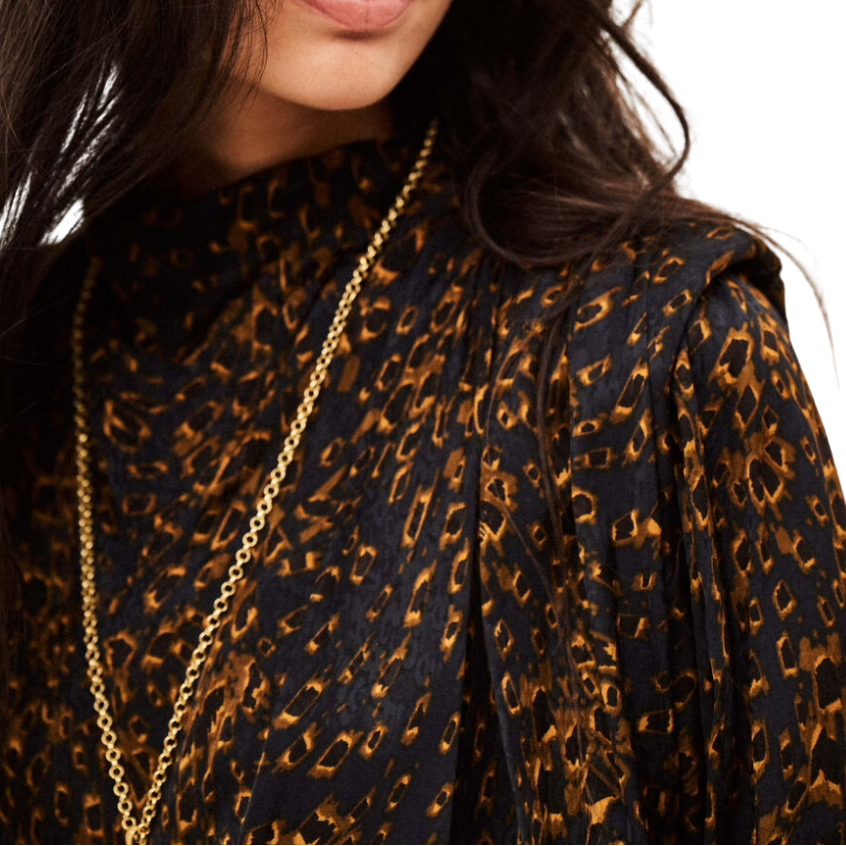 A close-up portrait of a woman wearing a black blouse with a brown leopard print pattern and a gold chain necklace, set against a plain white background.